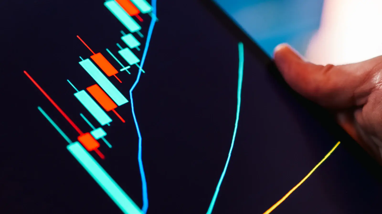 A hand holding a tablet showing a candlestick trading chart in the dark